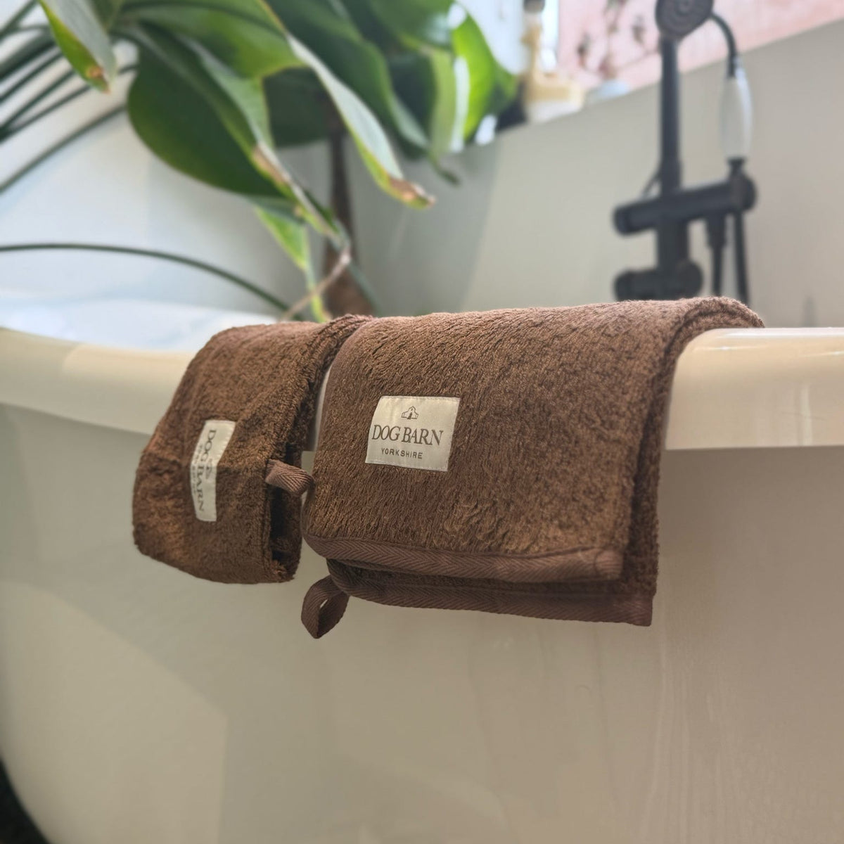 A brown plush cotton bath towel with a white 'DOG BURN' brand label, draped over the edge of a white freestanding bathtub with a black faucet and green plants in the background.