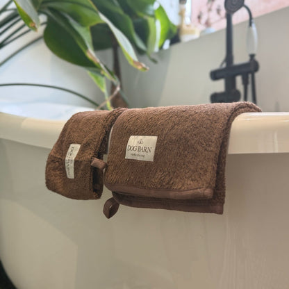 A brown plush cotton bath towel with a white 'DOG BURN' brand label, draped over the edge of a white freestanding bathtub with a black faucet and green plants in the background.