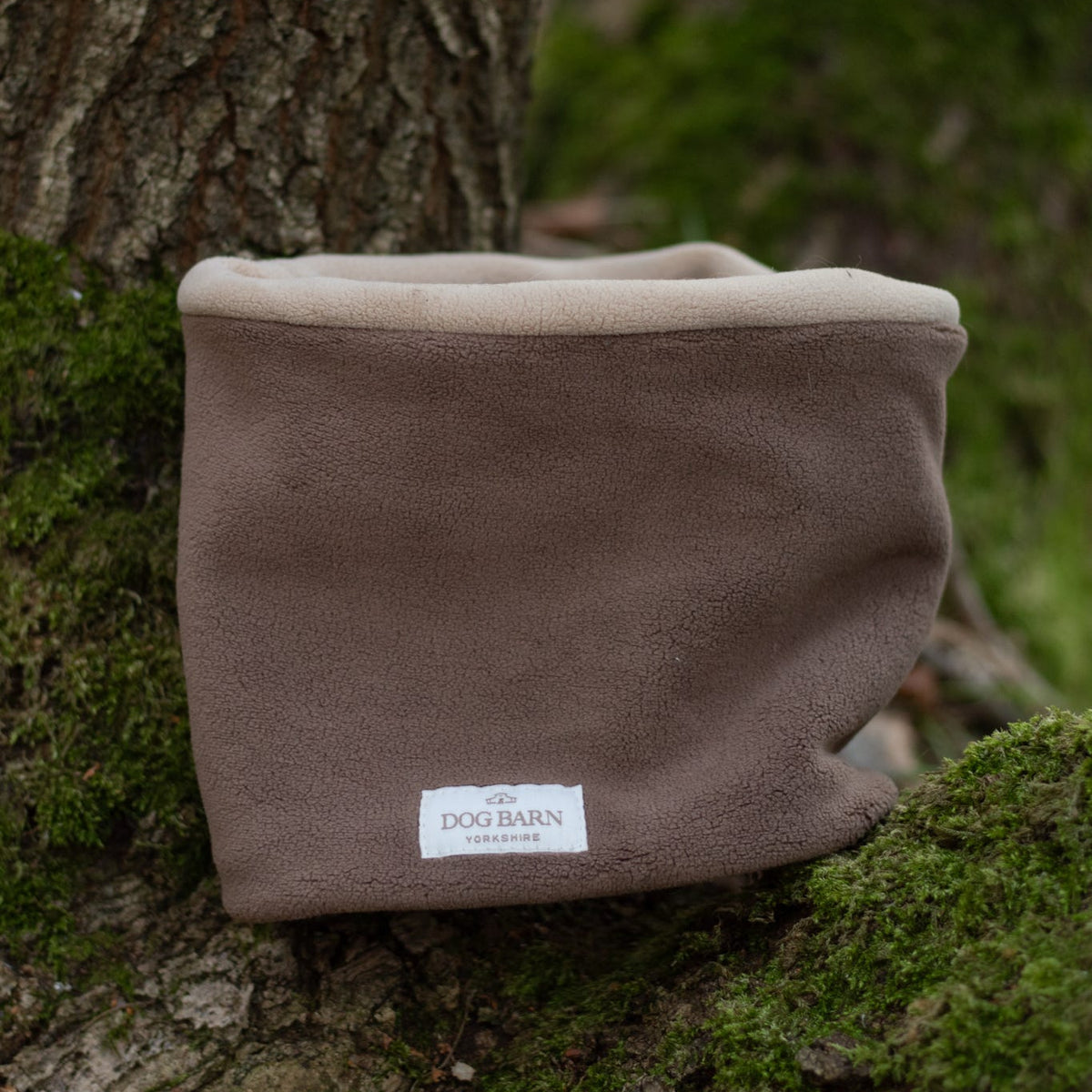 A brown fleece dog collar with a cream ribbed trim and a white 'DOG BARN INTERNATIONAL' label, resting on mossy tree bark outdoors.