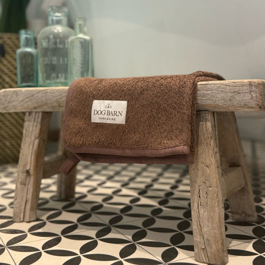 A brown plush bath towel with a white 'DOG BARN CLOSET' label, draped over a rustic wooden stool in a bathroom setting with a tiled floor and a bathtub in the background.