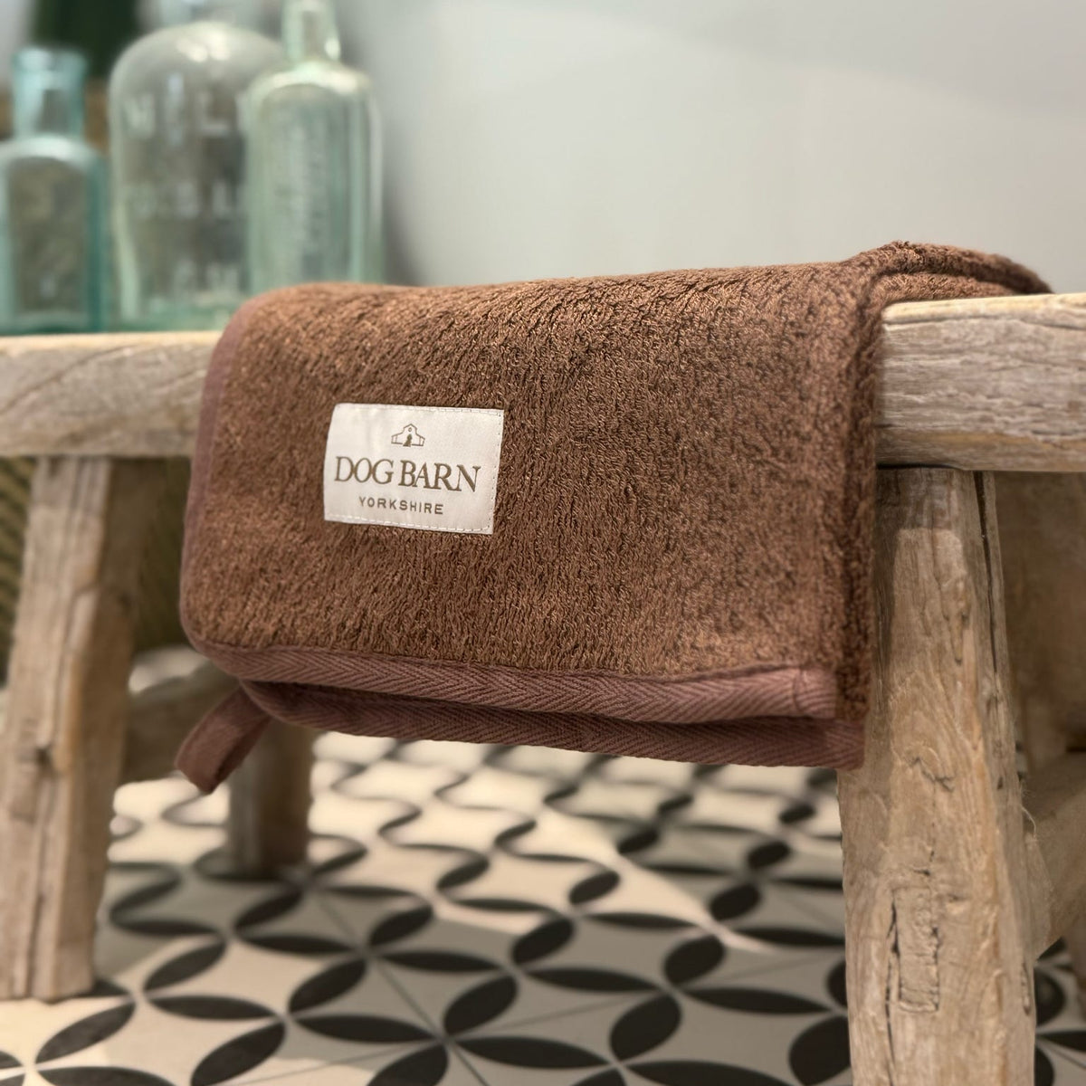 A brown textured pet towel with a white 'DOG BARN' label, draped over a rustic wooden bench in a home setting with patterned tile flooring and glass bottles in the background.